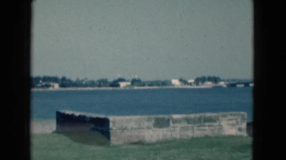 1975 Florida 8mm frame with grass, a low seawall, calm blue water, distant buildings, and soft film grain.