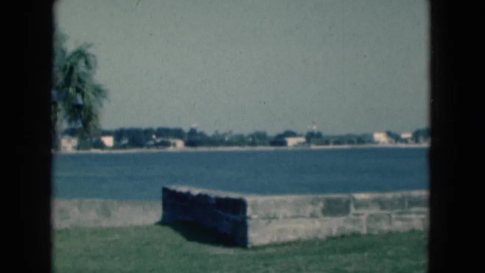 1975 Florida home-movie frame showing a park wall by blue water, distant shoreline buildings, hazy sky, and a partial palm frond.