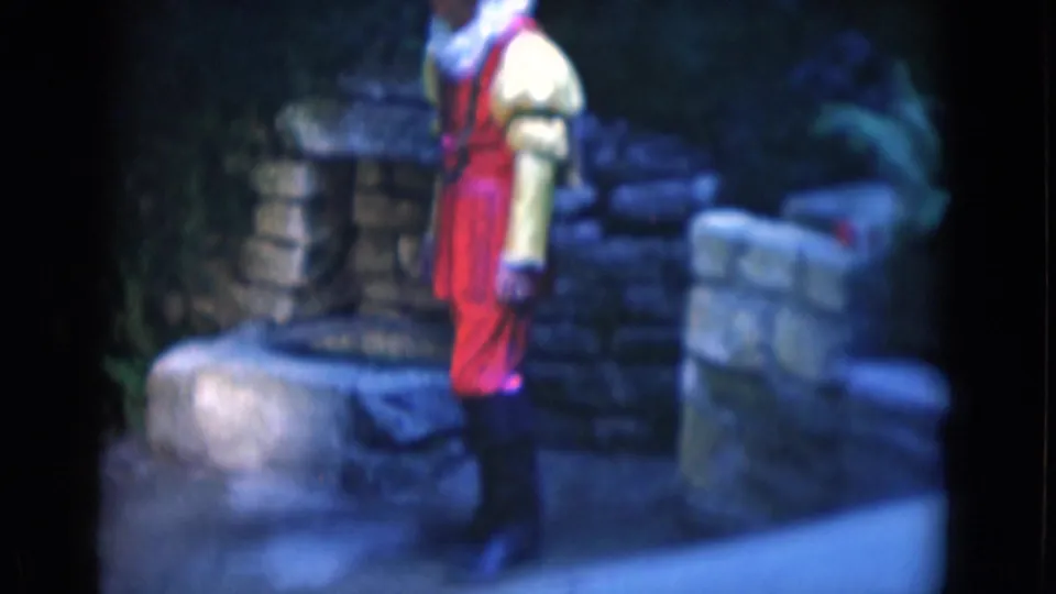 Soft 8mm view of the red and yellow costumed man standing by stone structures during a 1975 Florida visitor outing.