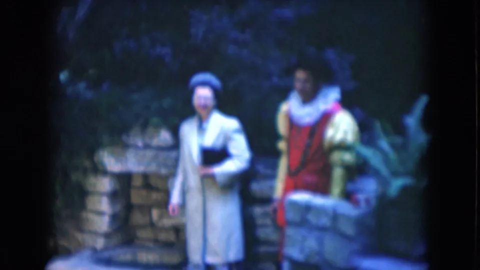 Woman in a light gray coat poses beside a red and yellow costumed man in a 1975 Florida outdoor attraction setting.