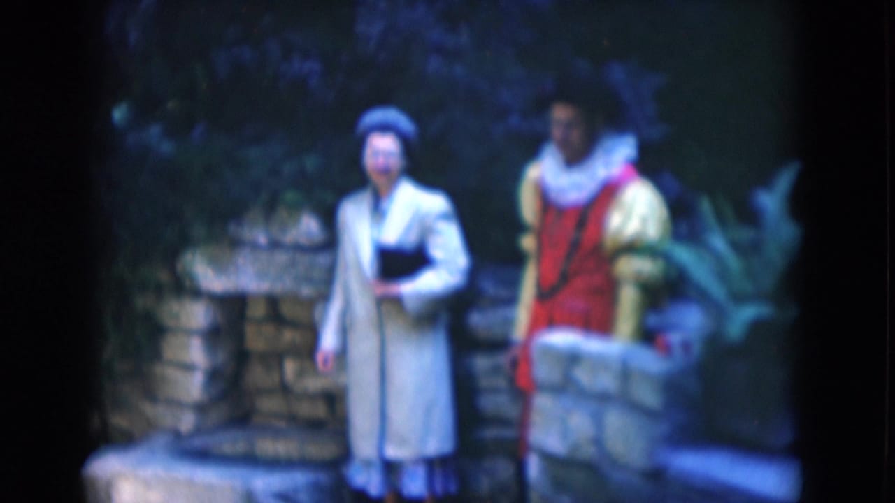In 1975 Florida 8mm footage, a woman in a pale coat and hat stands beside a young man in a red and yellow costume near stone steps and foliage.