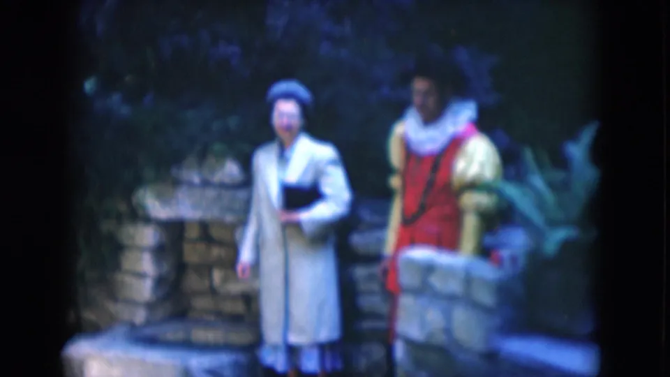 In 1975 Florida 8mm footage, a woman in a pale coat and hat stands beside a young man in a red and yellow costume near stone steps and foliage.