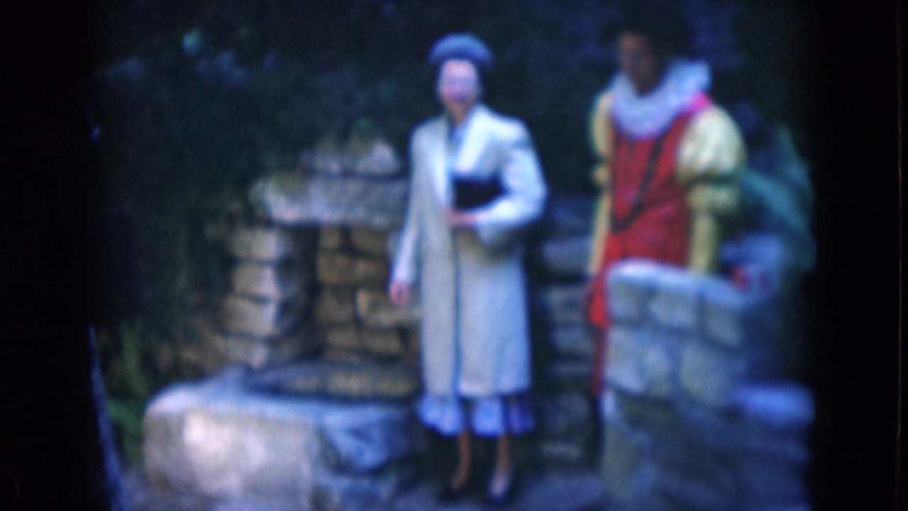 The costumed pair remains near stonework, with the woman holding a dark item and the young man standing by a low wall in soft 8mm color.
