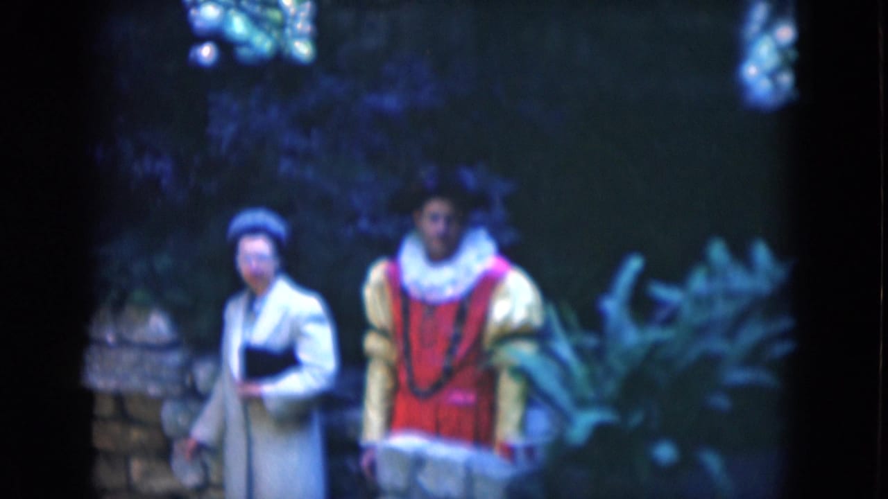 Two costumed adults walking beside stonework and foliage in a garden setting.