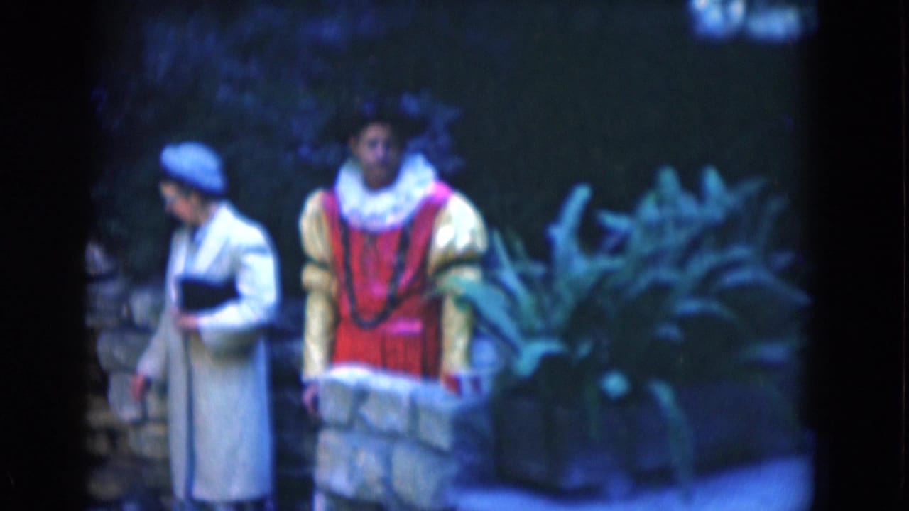 Two adults in historical-style costumes stand or move near a low stone garden wall in 1975 Florida footage.