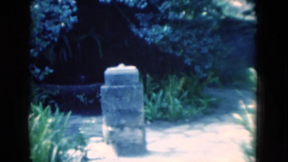 A small stone garden monument appears on a path surrounded by shrubs and foliage in the Florida home-movie scene.