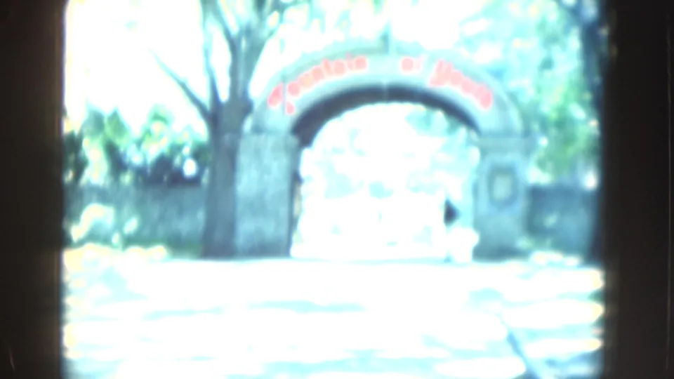 Overexposed 1975 Florida 8mm frame of a stone entrance arch with red lettering, trees, bright pavement, and a small person near the gateway.