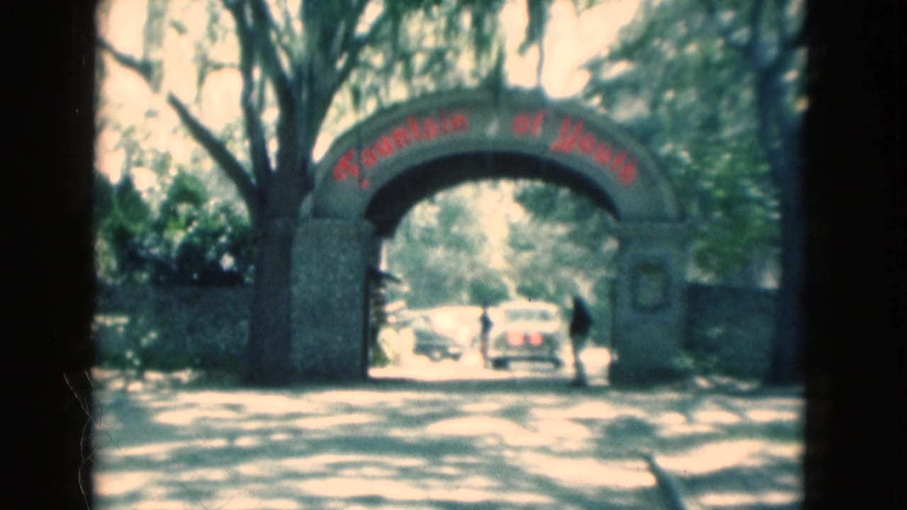 1975 Florida 8mm frame showing the Fountain of Youth entrance arch under trees, with cars beyond it and a person standing near the roadway.