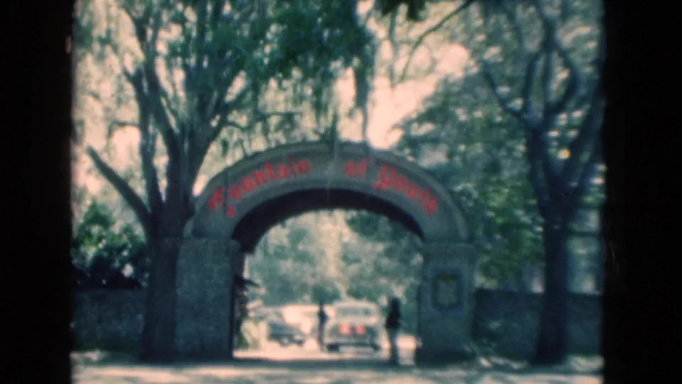 1975 Florida home-movie frame of the Fountain of Youth arch, partly softened by film blur, with vehicles beneath the arch and trees overhead.