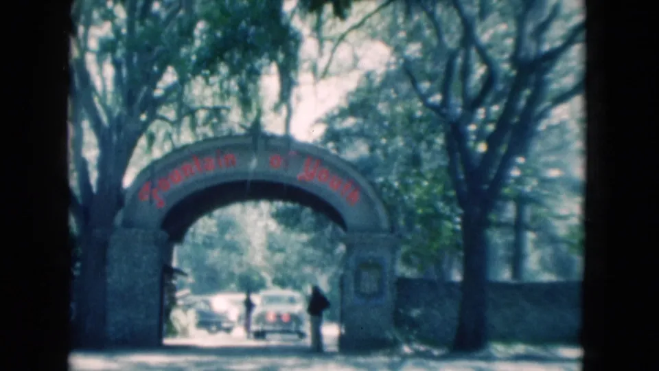 1975 Florida 8mm frame showing the Fountain of Youth entrance arch, red lettering, cars under the arch, and a person standing at the right side.