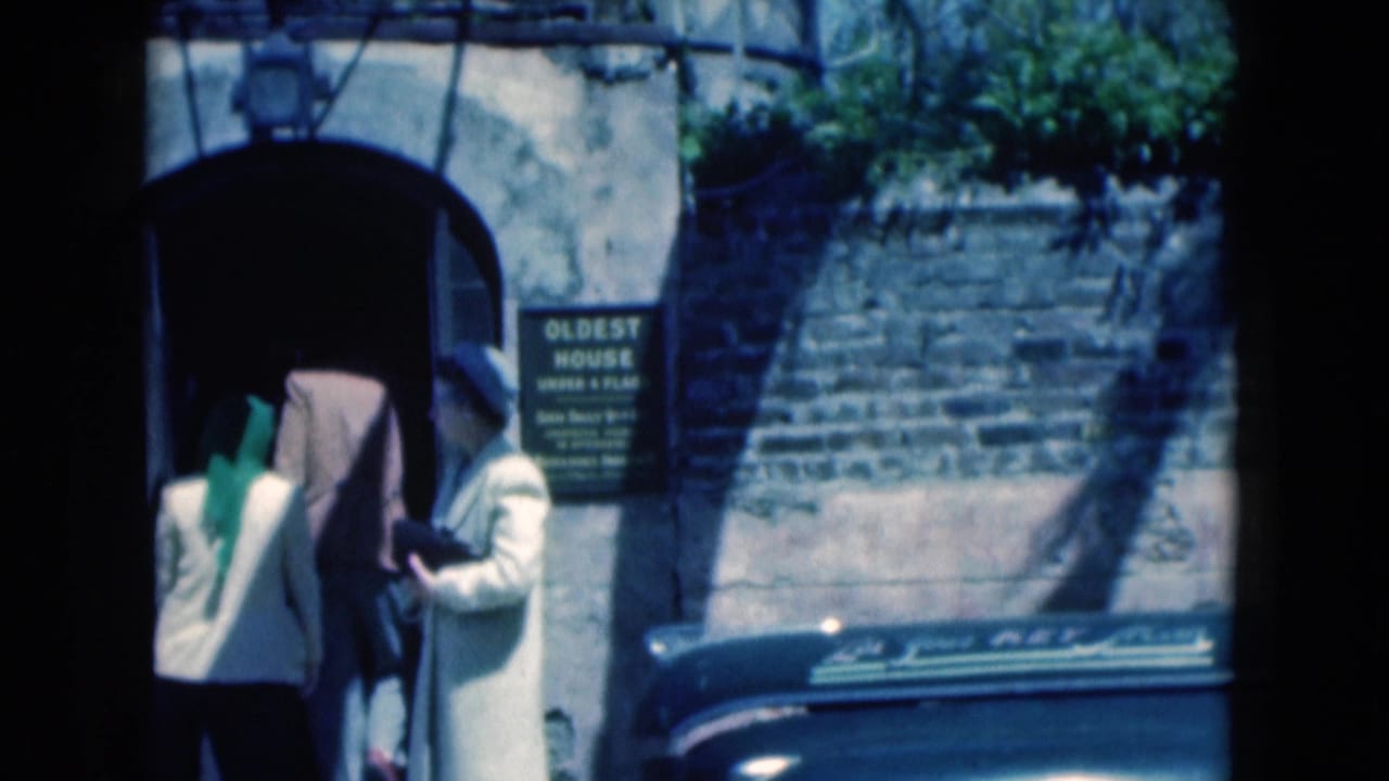 1975 Florida home-movie frame with two visitors entering a side doorway and a woman holding a book near the "OLDEST HOUSE" sign.