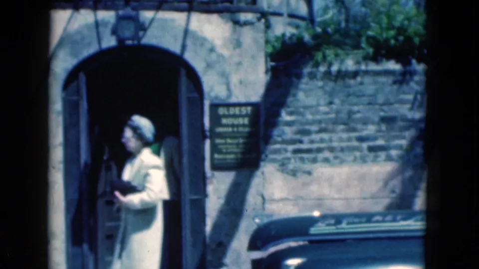 1975 Florida 8mm frame showing a woman in a light coat and hat at an arched doorway beside an "OLDEST HOUSE" sign.