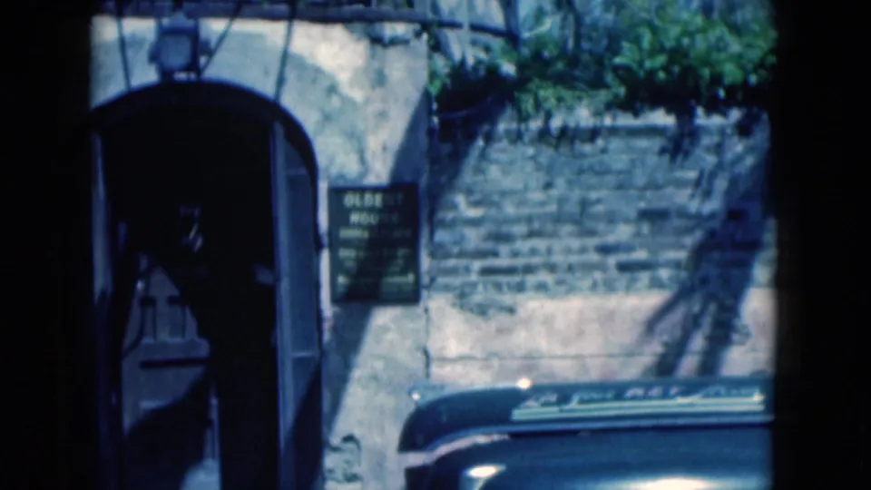 1975 Florida home-movie frame with the arched entrance, readable "OLDEST HOUSE" sign, stone wall, and a shadowed departing figure.