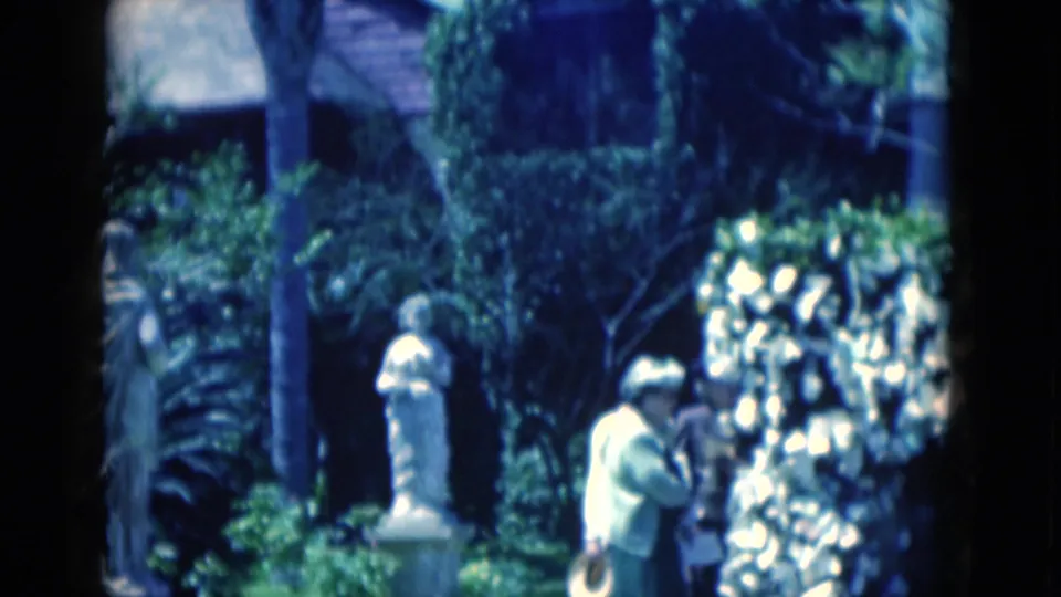 Visitors in light clothing stand beside a white statue and hedge in a 1975 Florida garden, with dense foliage and a building behind them.