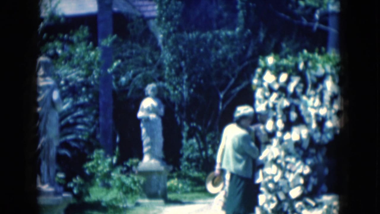 Two elderly women stand near white garden statues and dense greenery.