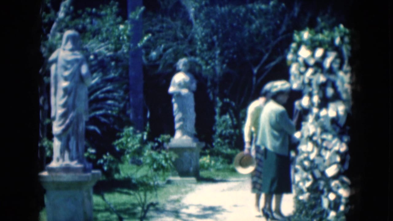 Two elderly women walk away along a garden path with white classical statues, greenery, and a decorative stone wall visible.