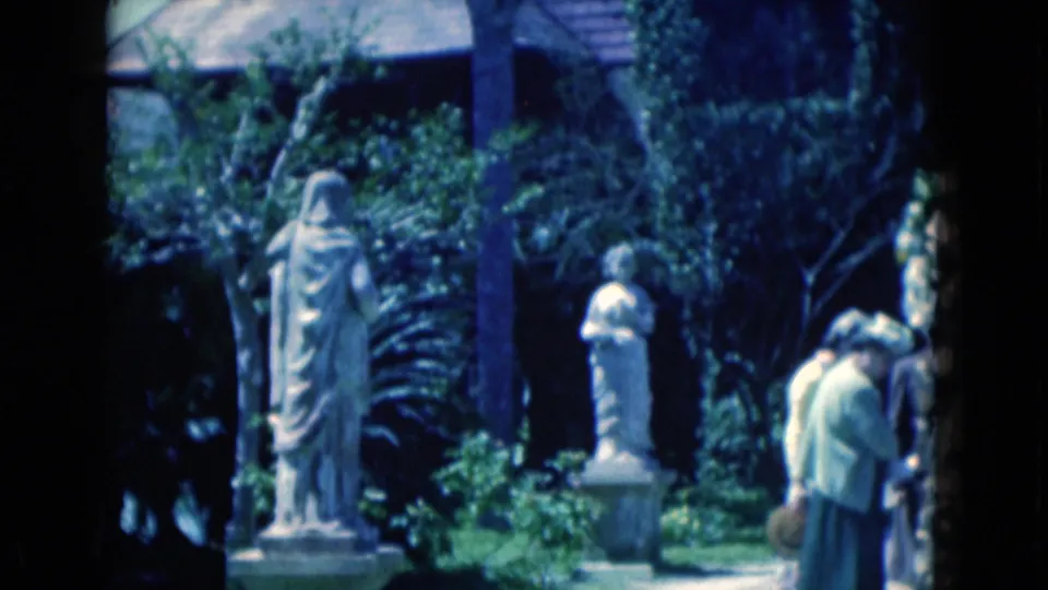A tall robed statue and smaller pedestal statue stand among shrubs as the two women pass partly out of frame in the garden.