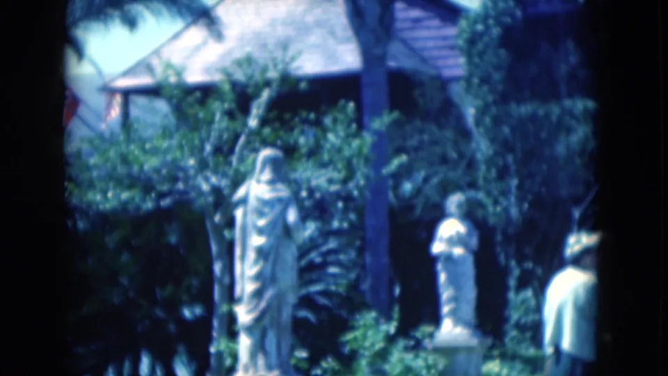 White classical statues, thick garden foliage, and a roofed structure fill the frame as one elderly woman moves out at the right edge.