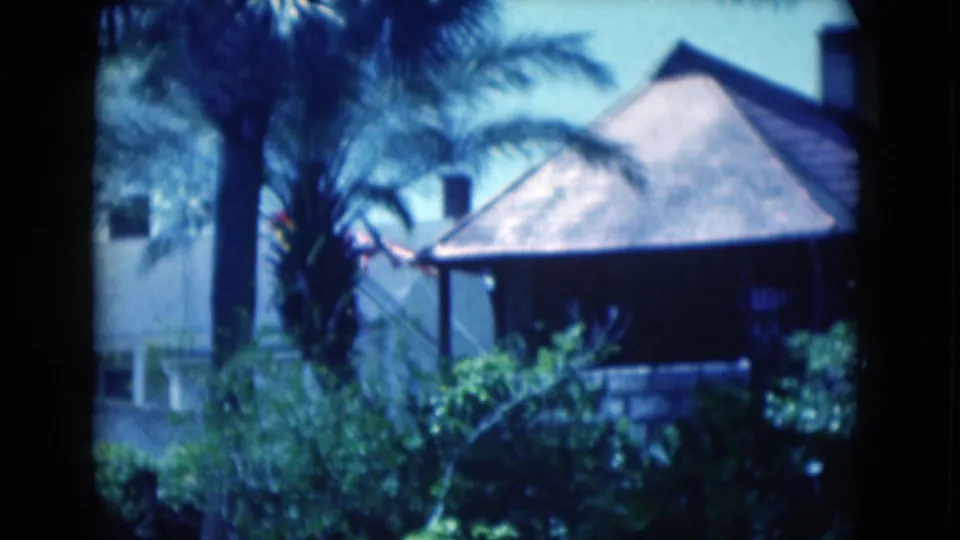 1975 Florida 8mm frame showing palms, shrubs, and a tile-roofed residence, with a possible distant person at left.