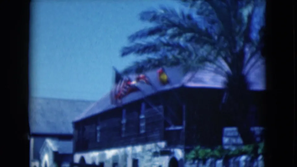 1975 Florida 8mm frame showing flags blowing above a dark exterior building, with palm fronds against a clear blue sky.