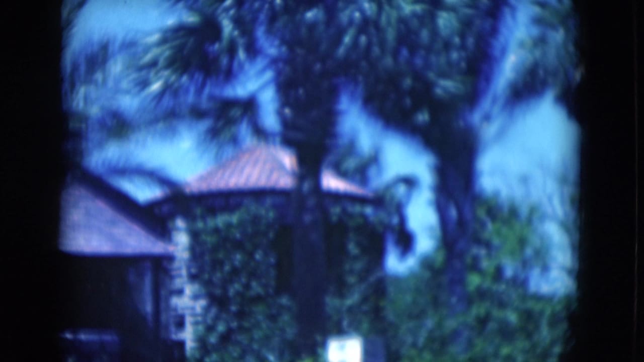 1975 Florida home-movie frame showing a tropical stone residence with red tile roof, palms, and a low boundary wall.