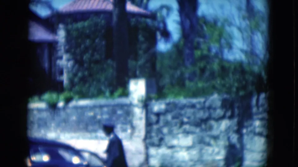 A distant person passes a parked car in front of the tropical Florida residence and low stone wall.