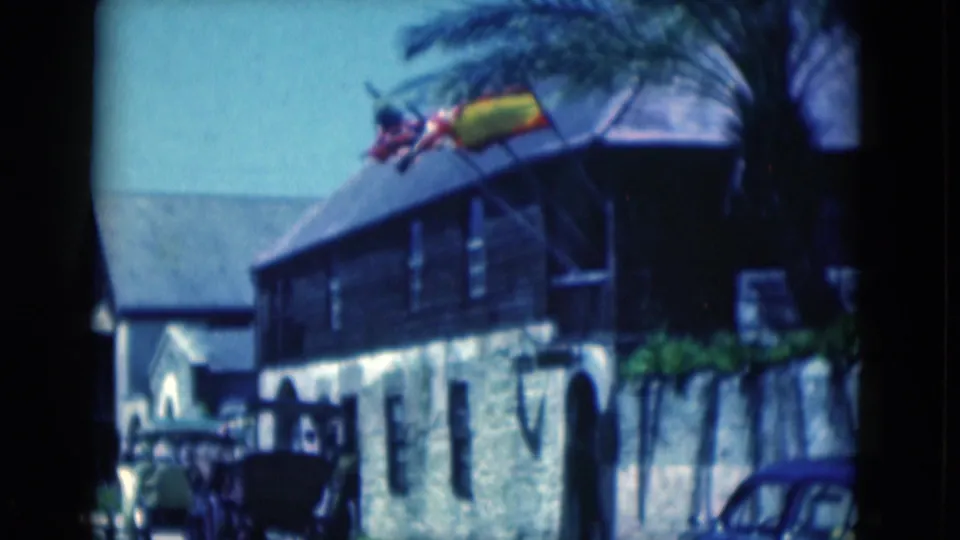 1975 Florida 8mm view down the street beside a stone-and-wood building with flags, a sidewalk, parked vehicles, and a blue car.
