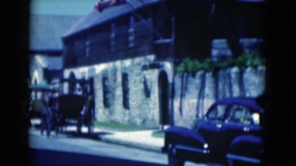 1975 Florida street frame showing the flagged stone-and-wood building, blue car, sidewalk, and a few distant pedestrians.
