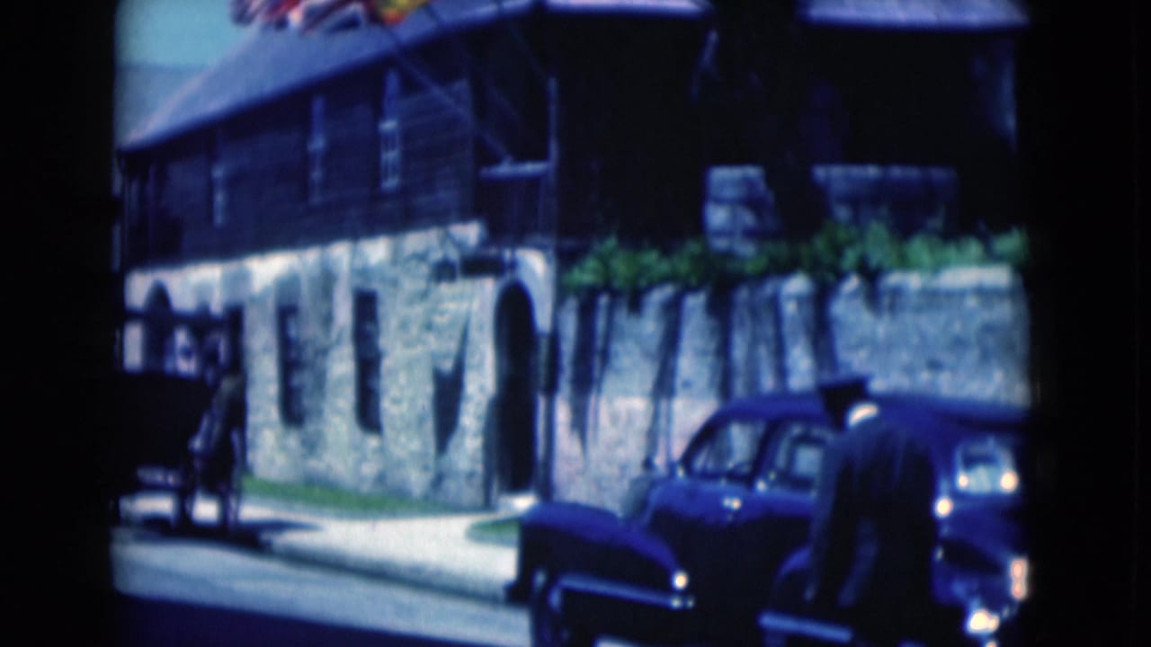 1975 Florida home-movie frame showing a man near a blue classic car outside a stone-and-wood building with flags above.
