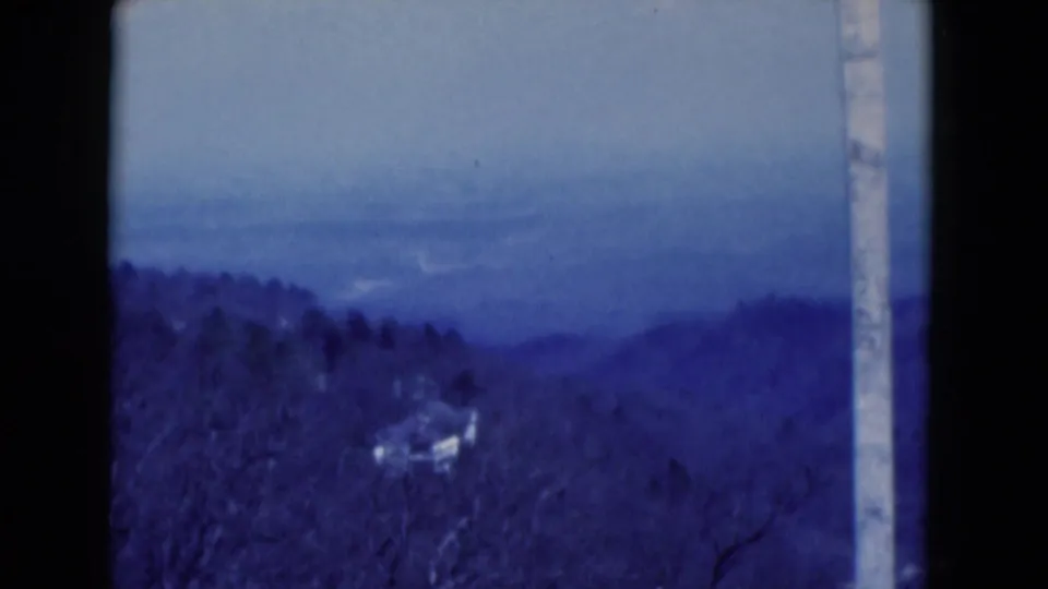 Reported 1975 Florida overlook view with wooded slopes, blue haze, and a pale railing post at the frame edge.