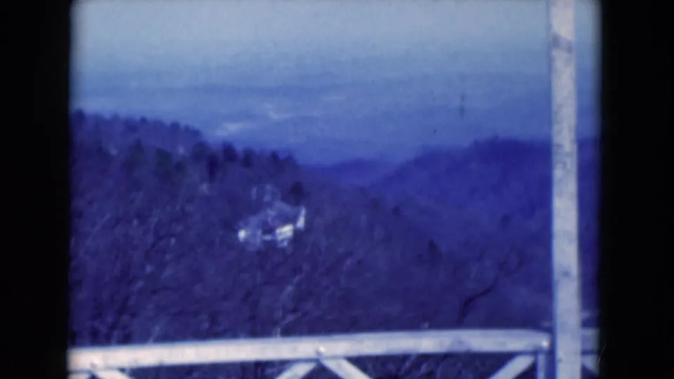 1975 8mm scenic overlook frame showing a wooden railing, wooded hills, and a small distant structure in blue-toned film.