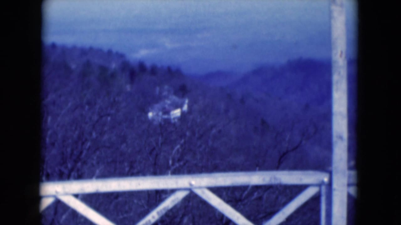 1975 8mm home-movie view of hazy hills beyond a white railing, framed by bare trees and a small distant hillside structure.