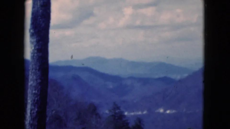 1975 8mm frame cataloged to Florida showing layered blue ridges, a cloudy sky, dark tree trunks, and distant pale highlights.