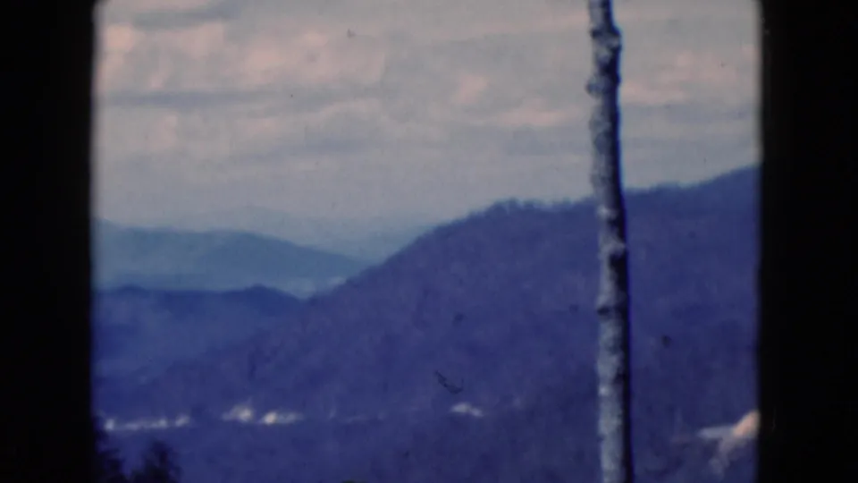 Final cataloged 1975 frame of the blue-tinted mountain vista, with layered ridges, cloud cover, film wear, and a vertical pole or trunk.