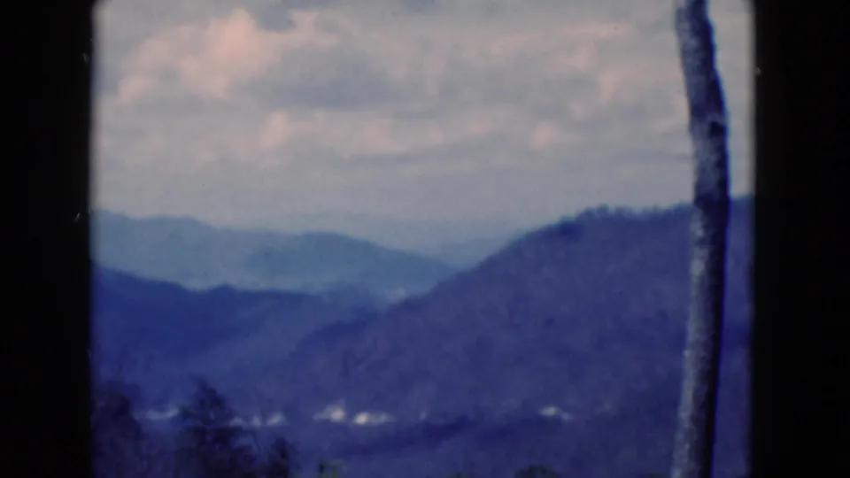 1975 8mm home-movie frame from Florida showing layered blue ridges, valley haze, cloudy sky, and a bare trunk near the right edge.