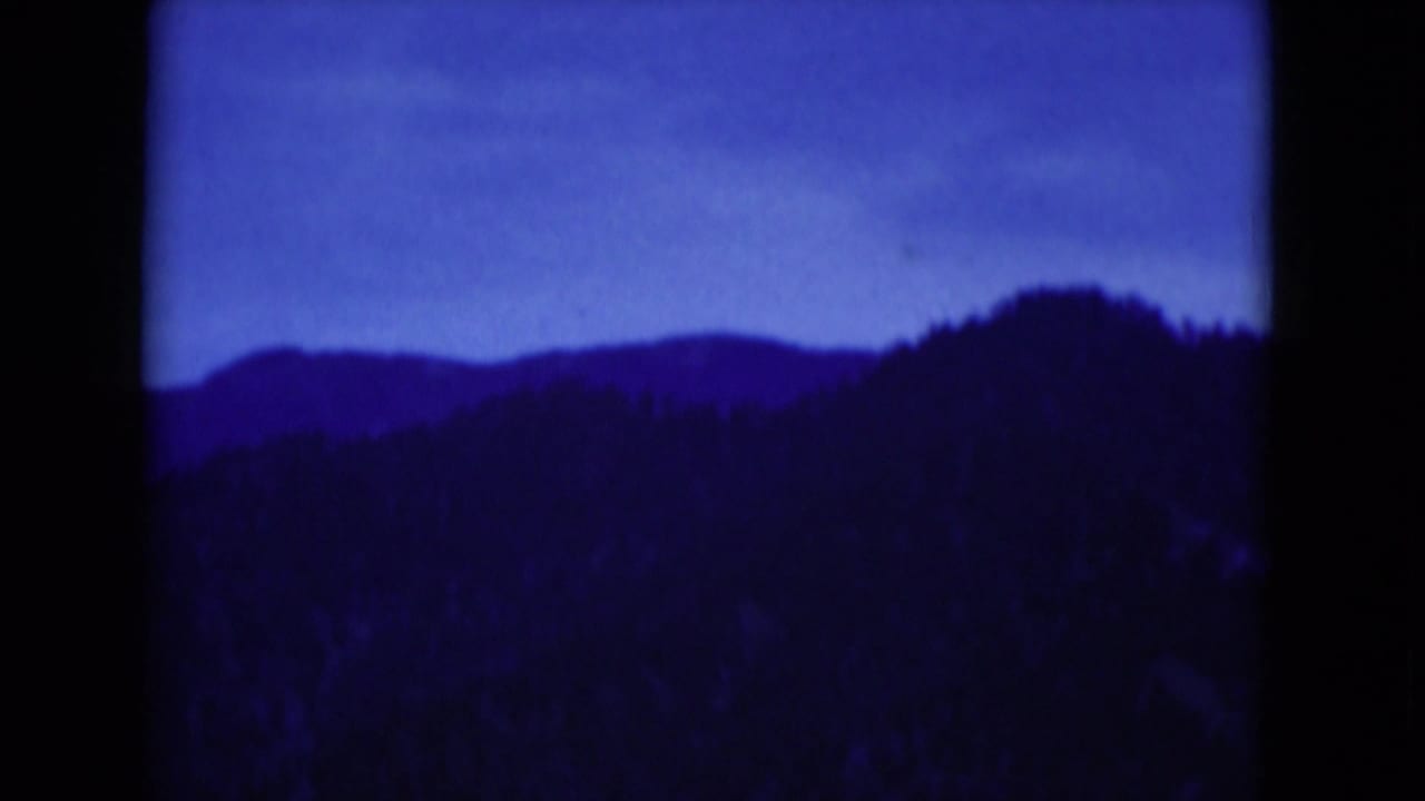 Blue-shifted 1975 8mm view of shadowed mountain silhouettes and wooded slopes, with dark film borders around the frame.