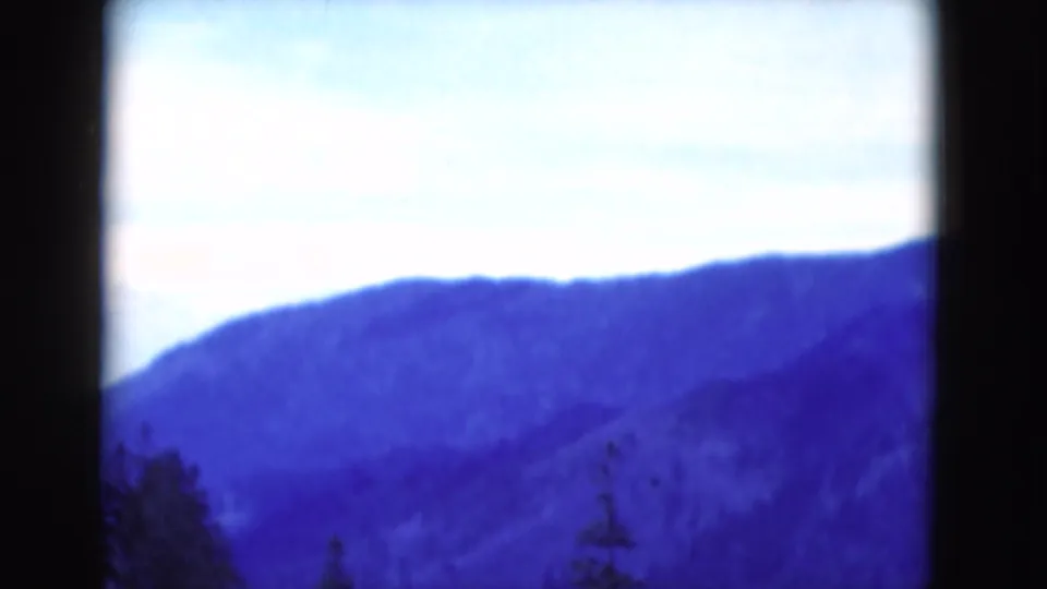 1975 8mm scenic frame with a bright pale sky, blue-tinted mountain ridge, and dark forested foreground.