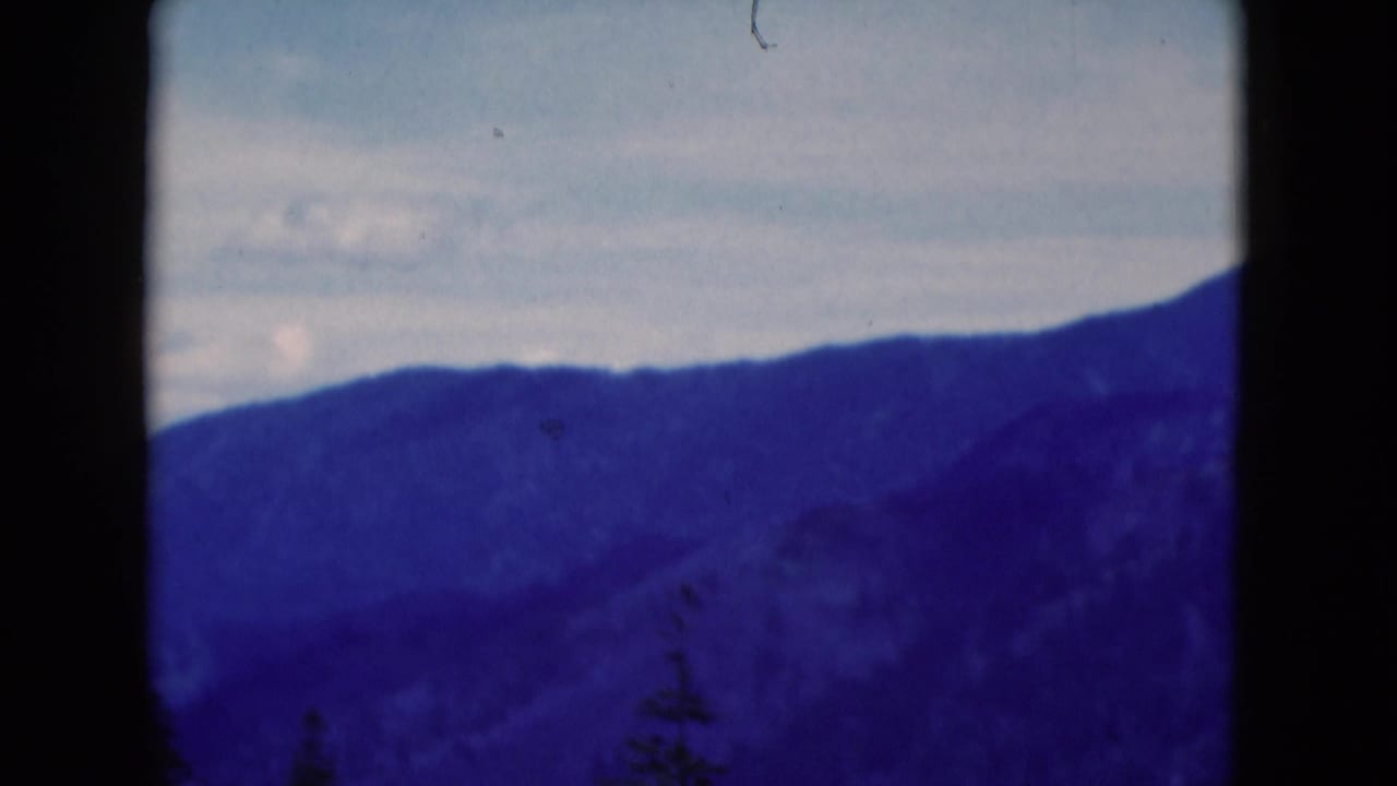 1975 8mm home-movie frame of a distant wooded mountain ridge under pale clouds, with dark film borders and blue-purple tint.