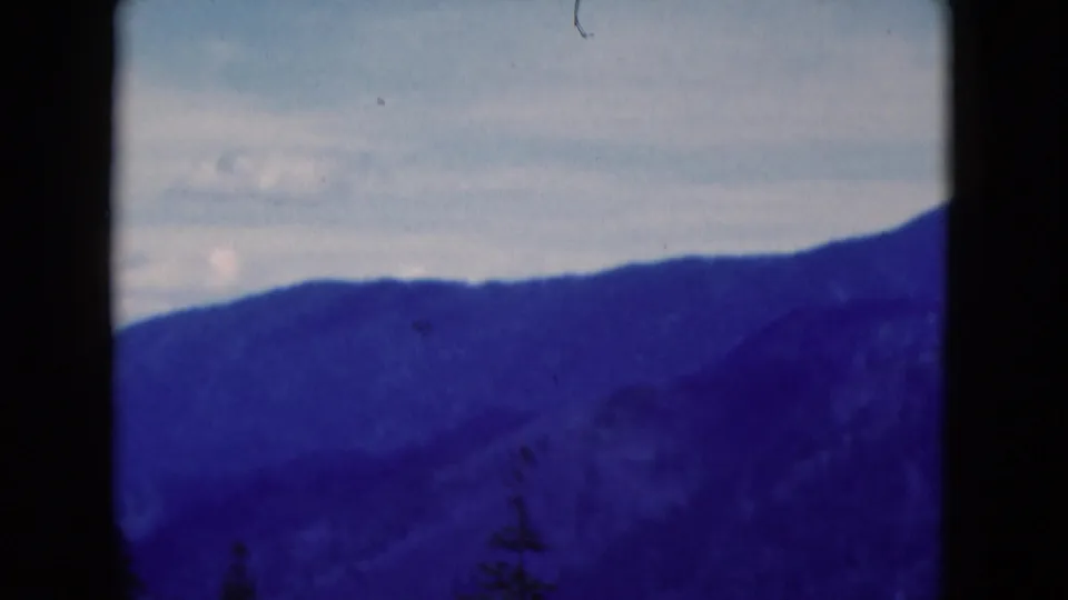 1975 8mm home-movie frame of a distant wooded mountain ridge under pale clouds, with dark film borders and blue-purple tint.