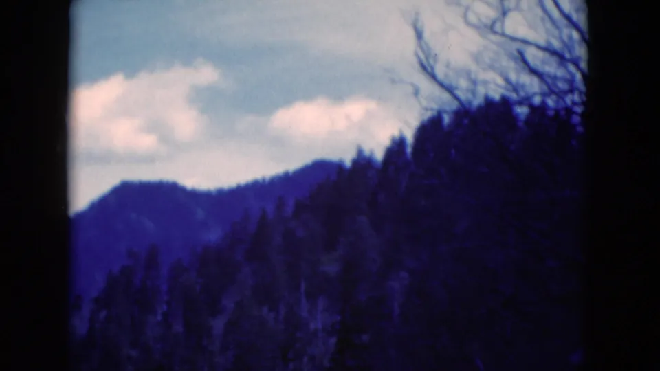 1975 8mm home-movie frame showing blue-tinted ridges, dark pine-covered foreground, and soft clouds overhead.