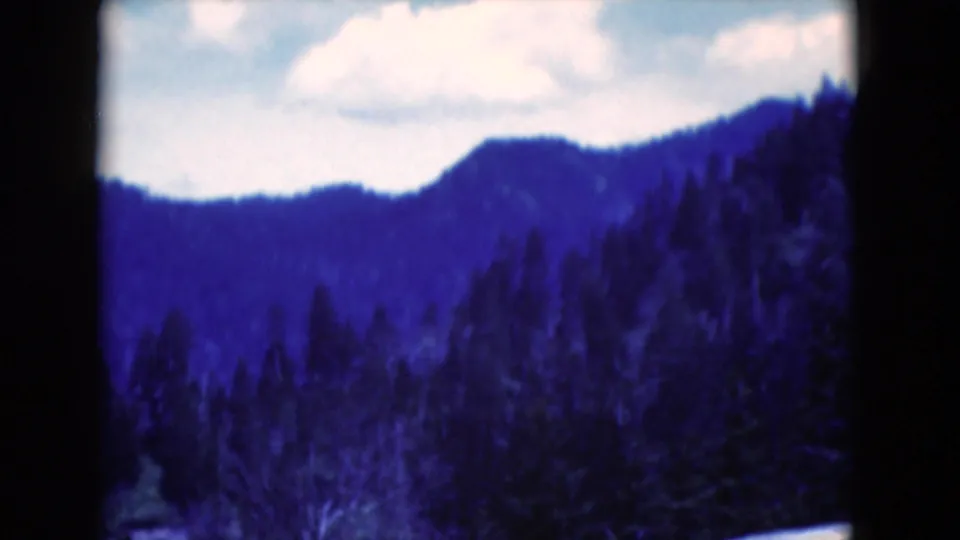 1975 8mm home-movie frame of a distant wooded hillside under pale sky, with a strong blue color cast and soft focus.