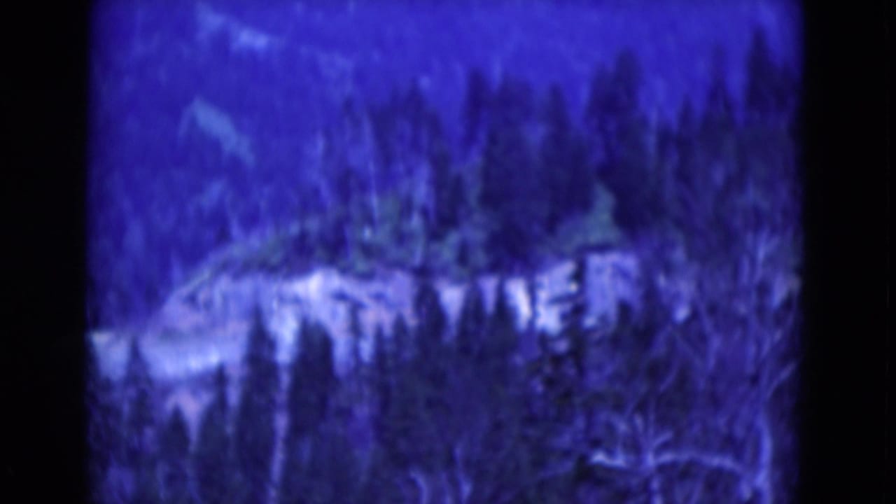Blue-shifted 1975 8mm home-movie frame showing trees and a pale rocky ridge or overlook area in a wooded mountain setting.