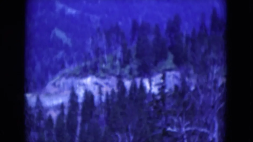 Blue-shifted 1975 8mm home-movie frame showing trees and a pale rocky ridge or overlook area in a wooded mountain setting.