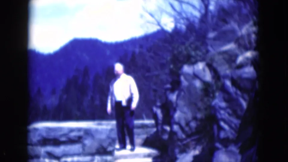 In 1975 8mm footage, an adult man stands on a stone overlook beside rocks, with dark wooded mountains and bright sky behind him.