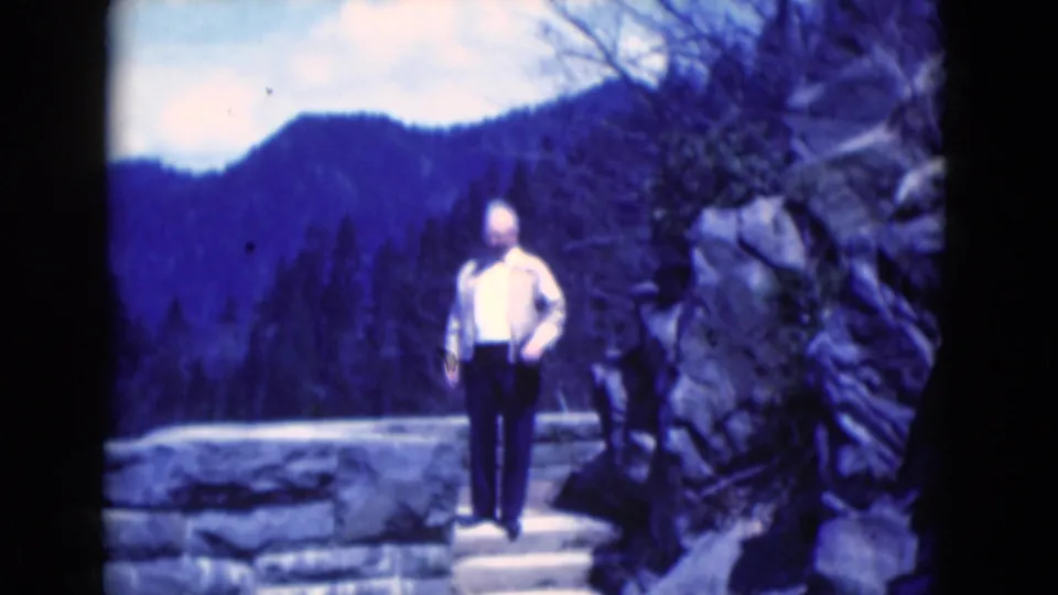 The man faces the camera area beside stonework and rocks, with blue-tinted wooded mountains behind him in the 1975 Florida clip.