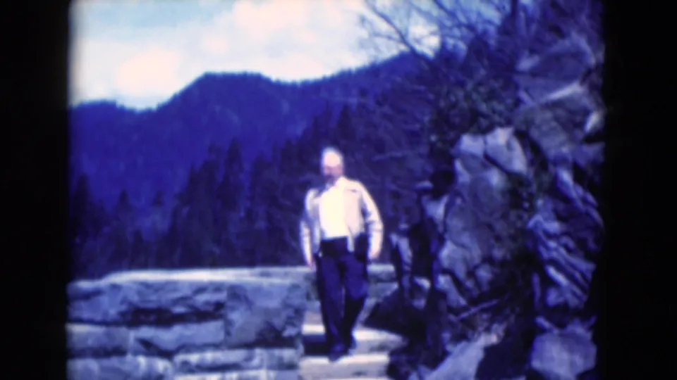 In the home-movie frame, the man descends short stone steps from the overlook, framed by rocks and distant wooded mountains.