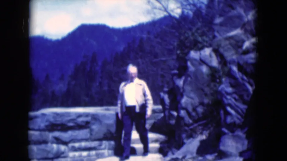 The man stands lower on the steps near the stone wall and rock face, with the blue-purple mountain landscape still visible behind him.