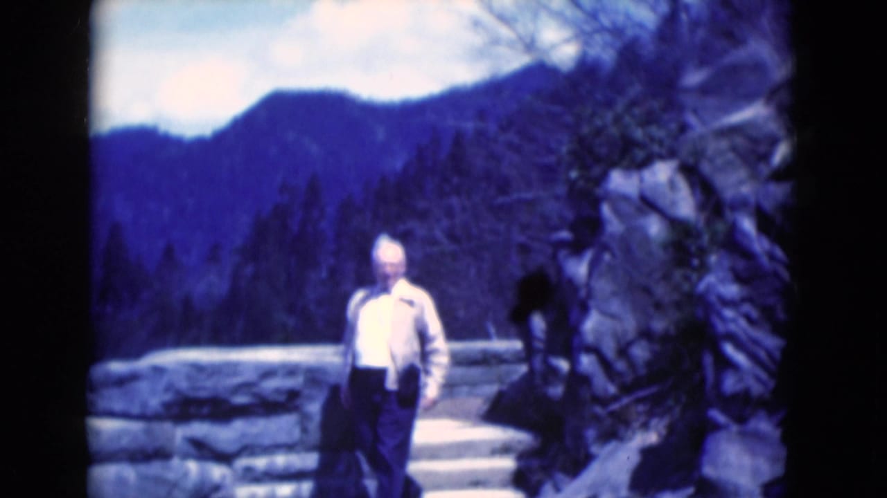 Man in a light jacket walking on a stone path with trees and distant ridges behind him.