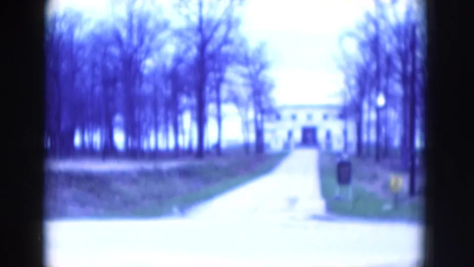 1970s 8mm frame showing a straight driveway through bare trees toward a pale building, with a small figure or object near the right side.