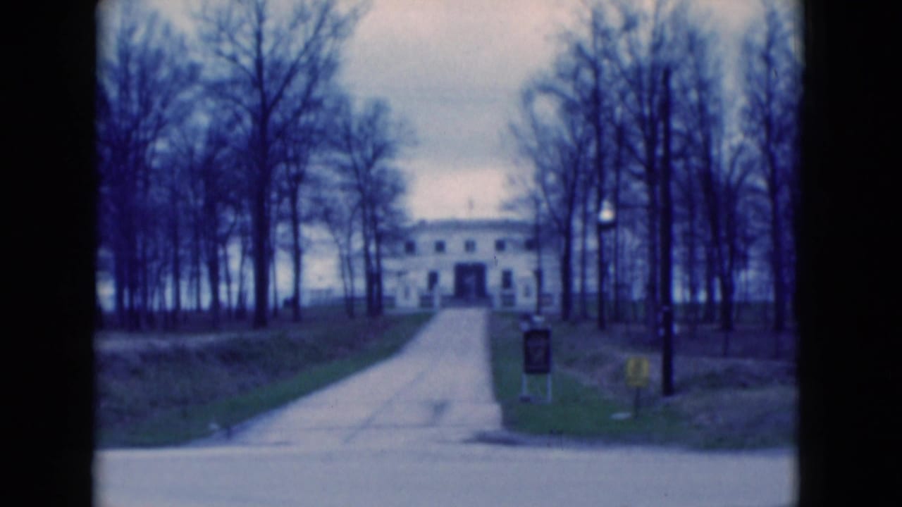 1975 Florida home-movie frame with a pale driveway rising toward a large white building, bordered by bare trees and small roadside signs.